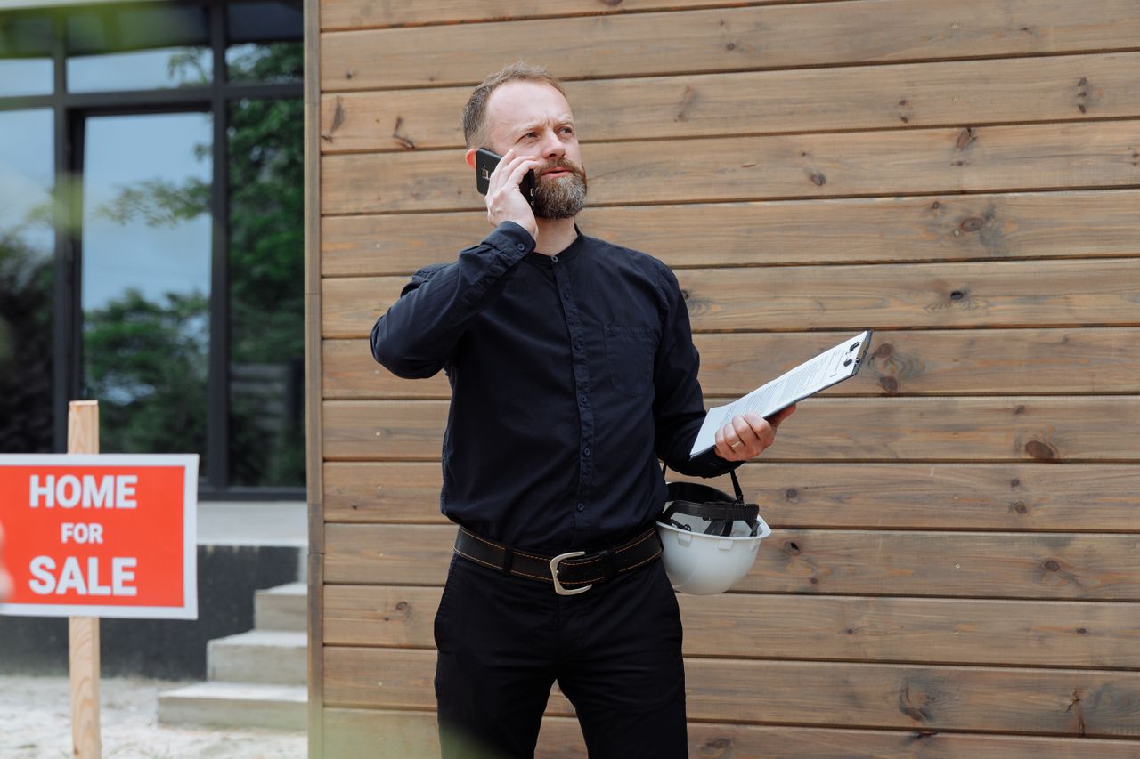 Home Real estate agent making a phone call while holding documents near home for sale sign.