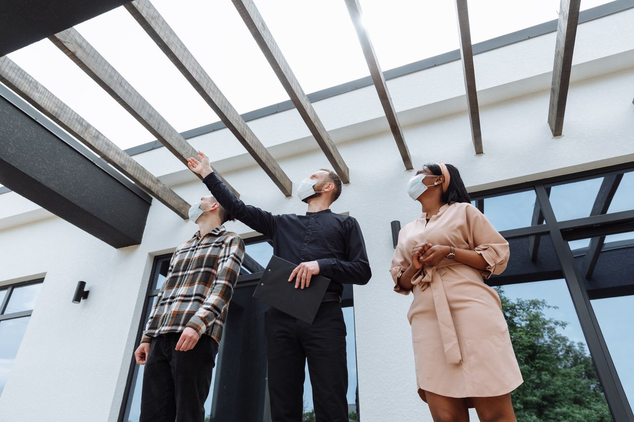 Masked real estate agent showcases modern architecture to clients outdoors.